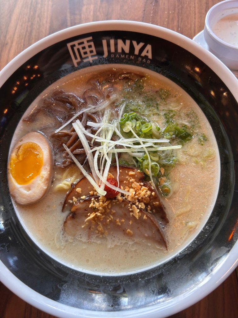 A bowl of ramen from Jinya Ramen Bar, featuring rich broth, soft-boiled egg, sliced pork, green onions, and crispy garlic on top.